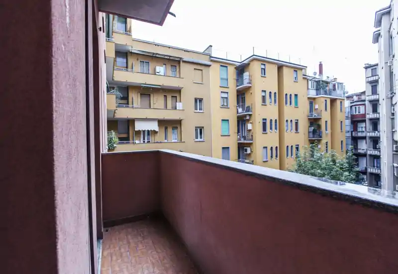 Piccolo balcone privato con vista sui palazzi vicini, pavimento piastrellato e parapetto a mezza altezza — ideale per un caffè mattutino o poche piante.