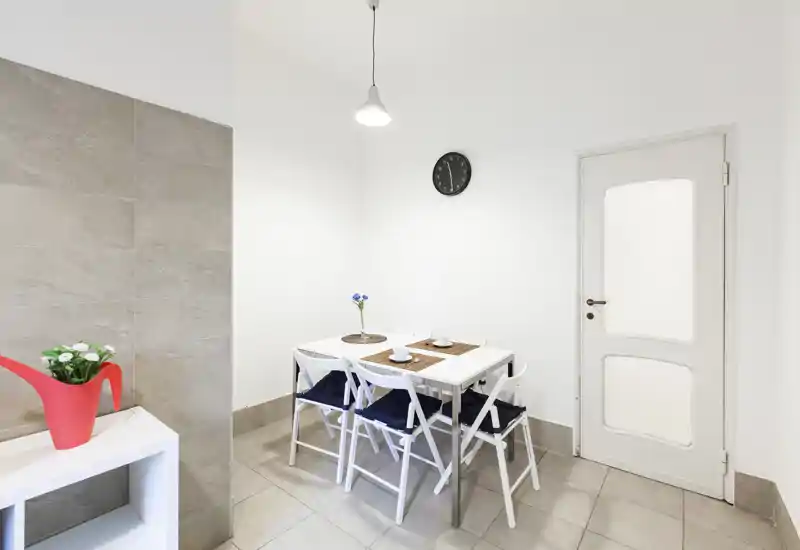 Angolo pranzo moderno e minimale con tavolo bianco, quattro sedie pieghevoli con cuscini, tovagliette e un piccolo vaso decorativo — luminoso e pronto per i pasti.