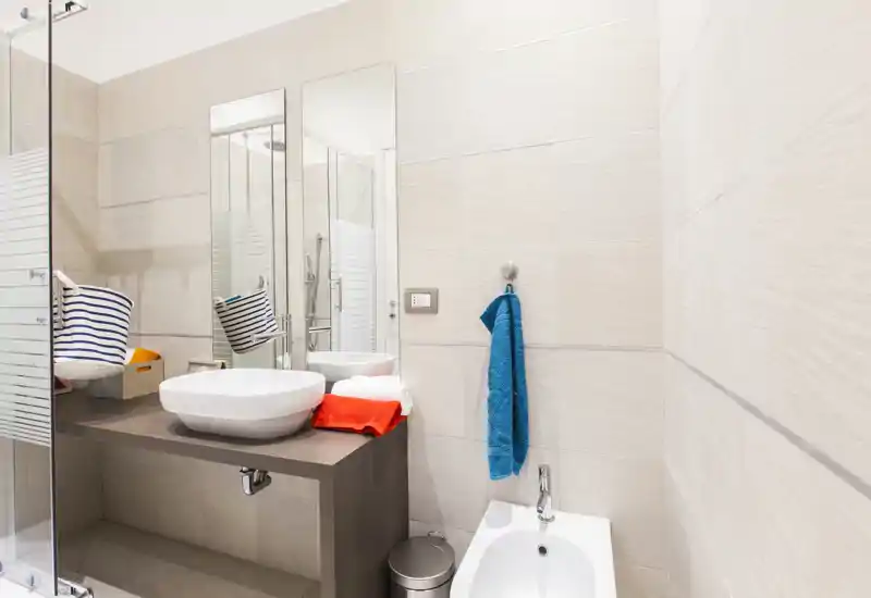 Bagno moderno e luminoso con lavabo a bacinella su piano, grande specchio, bidet e doccia in vetro visibili. Piastrelle neutre e asciugamani colorati creano un ambiente pulito e accogliente.