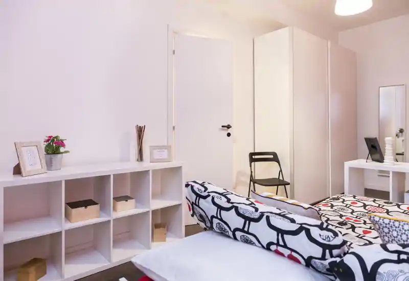 Camera da letto luminosa e ordinata con biancheria con motivo, cuscini e un armadio. La stanza è ben illuminata e allestita per foto di affitto, con un'atmosfera accogliente e spazio contenitivo pratico.