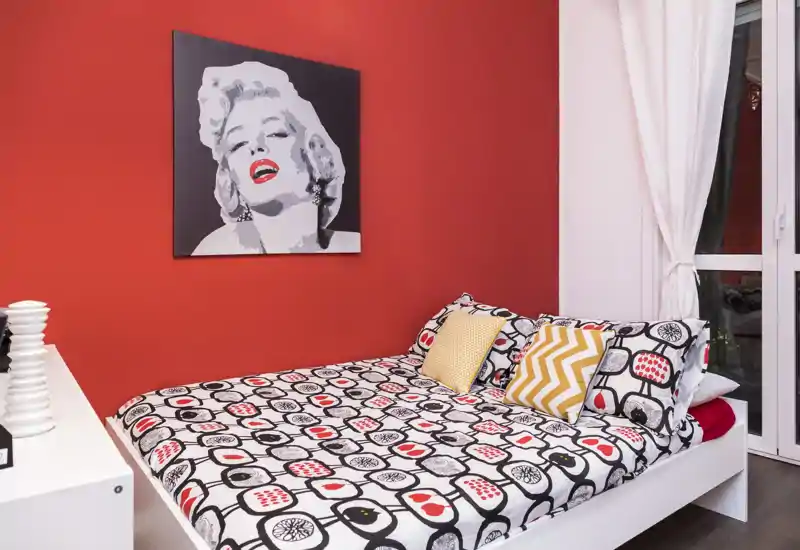 Camera da letto luminosa e di design con letto matrimoniale dal copriletto grafico su una parete rossa a contrasto e cuscini decorativi; ampia finestra con tende.