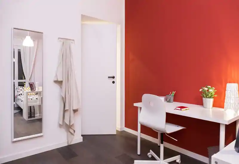 Rincón de dormitorio bien iluminado con escritorio blanco y silla frente a una pared roja; un espejo a la izquierda refleja la cama en la zona contigua, aportando profundidad.