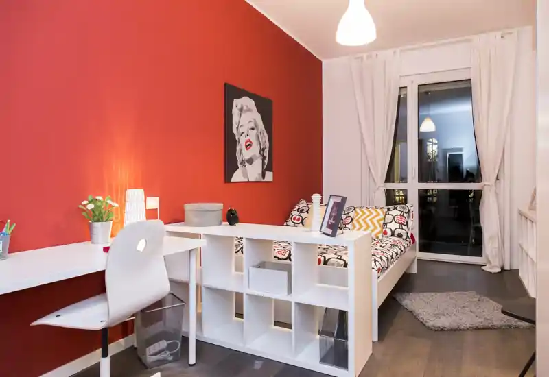 Camera da letto moderna e luminosa con letto bianco e cuscini decorativi su una parete d'accento rossa. Una scrivania compatta e scaffalatura offrono un comodo spazio di lavoro, le grandi finestre e l'illuminazione tenue rendono l'ambiente arioso e accogliente.