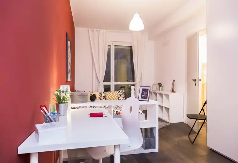 Dormitorio pequeño, luminoso y acogedor con una mesa blanca en primer plano y una cama/sofá cama ordenada junto a la ventana, pared roja de acento y cojines decorativos.