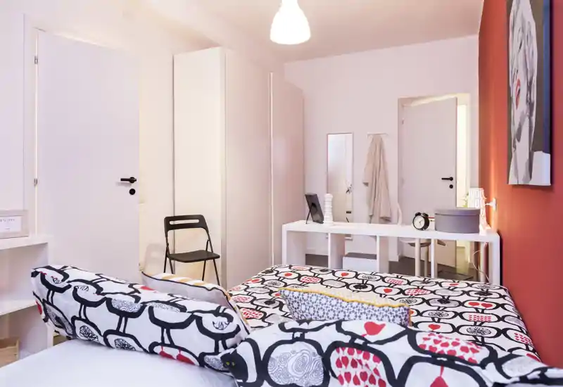 Camera da letto luminosa e ben allestita con biancheria a fantasia e una parete rossa. Il letto è il punto focale, affiancato da armadio, scrivania e specchio per un ambiente accogliente.