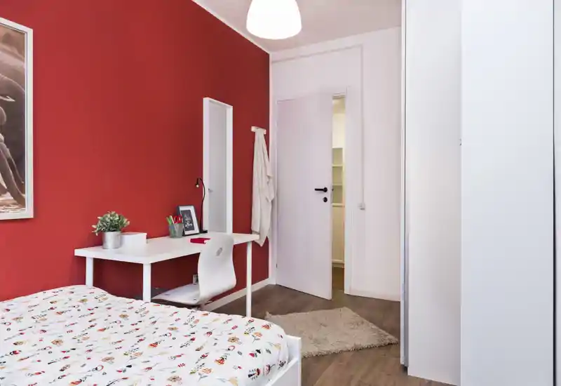 Camera da letto luminosa e accogliente con piumone a fantasia, scrivania bianca e sedia davanti a una parete rossa accesa; pavimento in legno e morbido tappeto completano l'ambiente moderno.