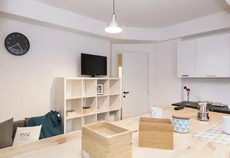 Zona de estar luminosa y moderna con televisor pequeño, estanterías abiertas y encimera de madera clara — cojines y decoración sencilla que crean un ambiente acogedor.