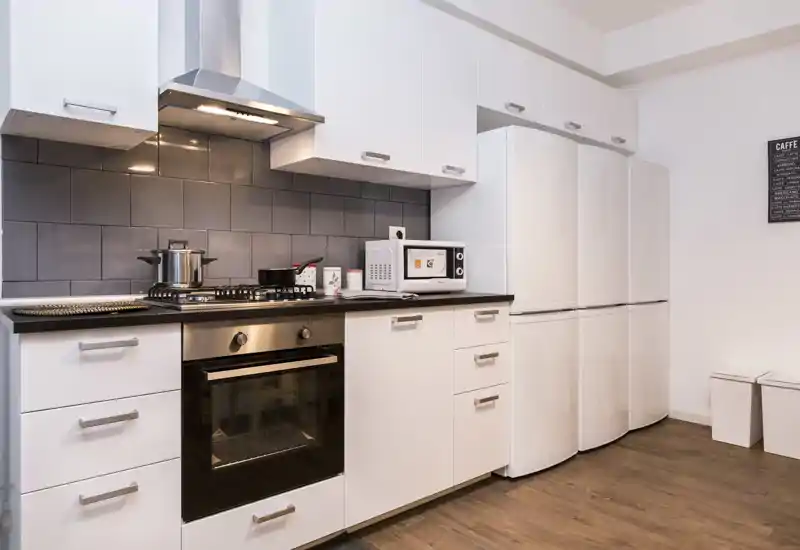 Cocina moderna y compacta con muebles blancos, horno empotrado, placa de gas, microondas y frigorífico alto; diseño limpio y minimalista con salpicadero de azulejos oscuros y suelo efecto madera.
