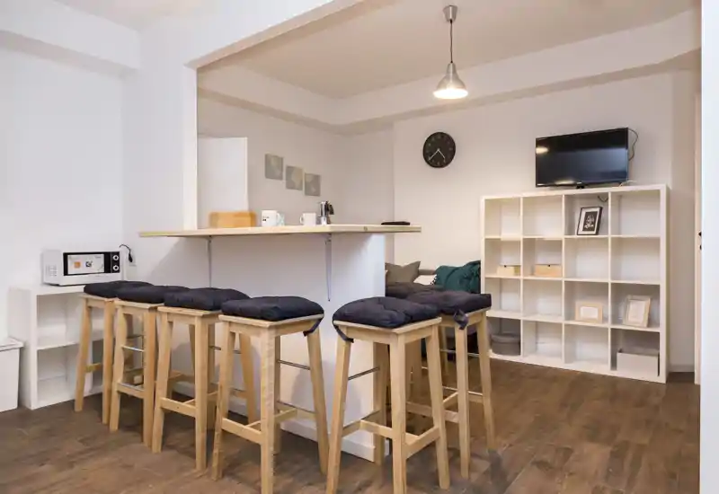 Área de estar moderna y luminosa con barra y taburetes altos, estantería modular y televisión en la pared: un espacio común compacto y confortable.