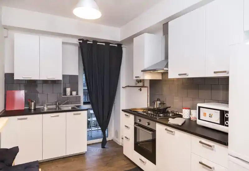 Cocina moderna y compacta con mobiliario blanco, placa de gas, horno integrado y microondas; encimera oscura y salpicadero de azulejos que ofrecen un espacio funcional con acceso al balcón.
