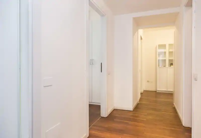 Pasillo luminoso y minimalista con suelo de madera cálida, paredes blancas y puertas que comunican con otras estancias — ideal para mostrar la circulación del piso.