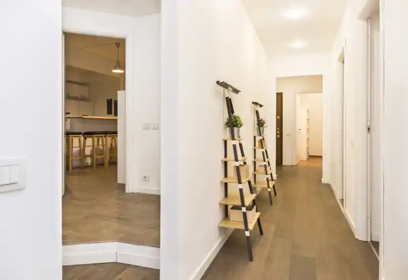 Entrada moderna y luminosa con suelo de madera, estanterías minimalistas en la pared y focos empotrados. El pasillo conecta con las habitaciones adyacentes y ofrece una primera impresión acogedora.