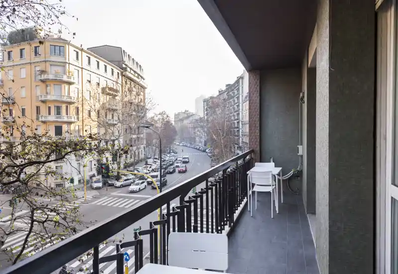 Balcón urbano soleado con suelo de baldosas, barandilla metálica y una pequeña mesa con sillas — ideal para tomar un café por la mañana contemplando la calle y los edificios históricos.