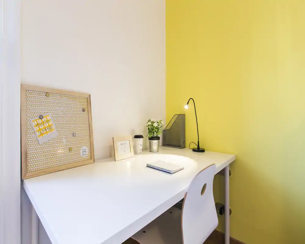 Angolo studio luminoso e ordinato con scrivania bianca, lampada da tavolo, piccola pianta e bacheca: perfetto per lavoro o studio da remoto.