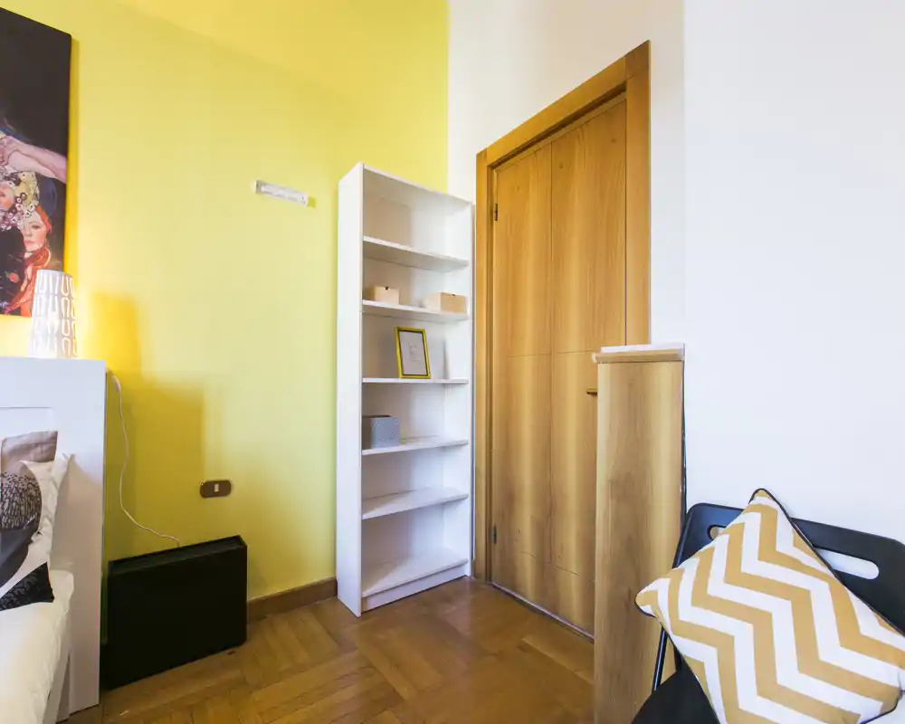 Angolo di camera luminoso e moderno con testiera visibile, cuscino a motivo e scaffale ordinato; parete gialla e porta in legno creano un’atmosfera calda.