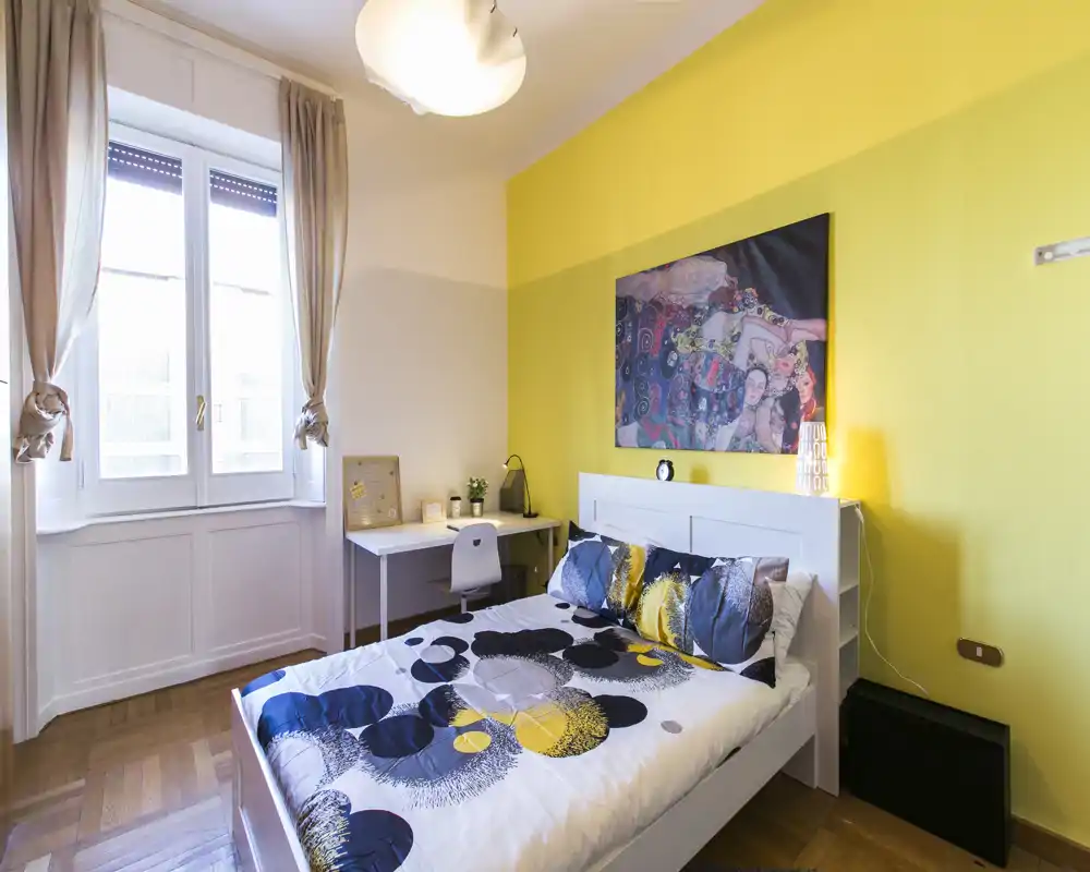 Camera da letto luminosa e curata, con letto matrimoniale, cuscini decorativi e ampia finestra che garantisce luce naturale; scrivania accanto alla finestra adatta per lavoro o studio.