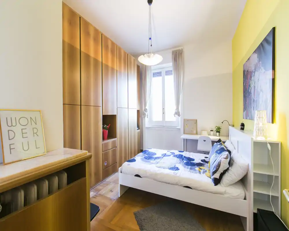 Camera da letto luminosa e ordinata con letto singolo, ampia armadiatura in legno, scrivania vicino alla finestra e pavimento in parquet — ambiente accogliente e ben illuminato ideale per una persona.