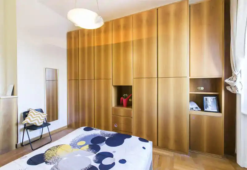 Camera da letto luminosa con letto matrimoniale a fantasia, ampie armadiature a muro in legno e luce naturale calda dalla finestra — moderna e ordinata.