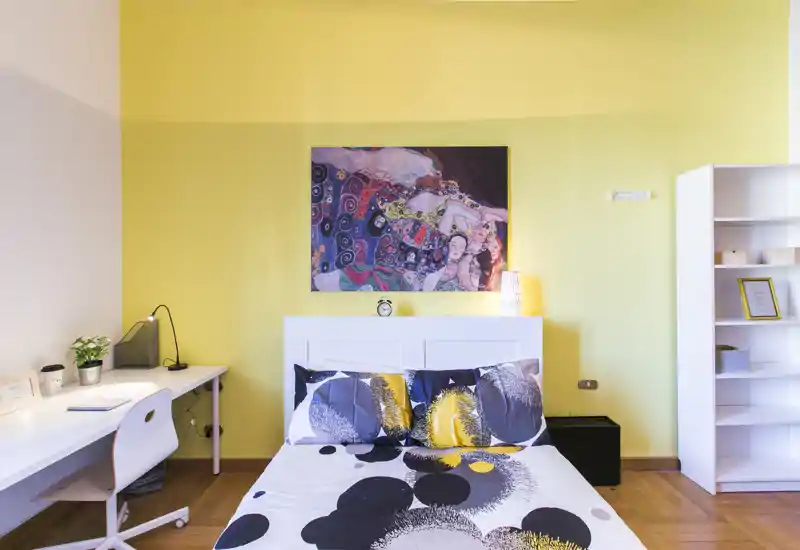 Camera da letto luminosa e di stile con letto matrimoniale al centro, cuscini decorativi e quadri moderni; una scrivania ordinata e scaffalatura completano l'ambiente.