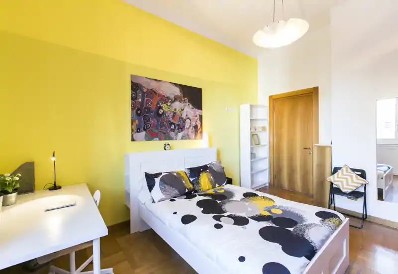 Camera da letto luminosa e di design con letto matrimoniale, biancheria decorata, scrivania e parete gialla a contrasto; ben illuminata e curata per annunci.