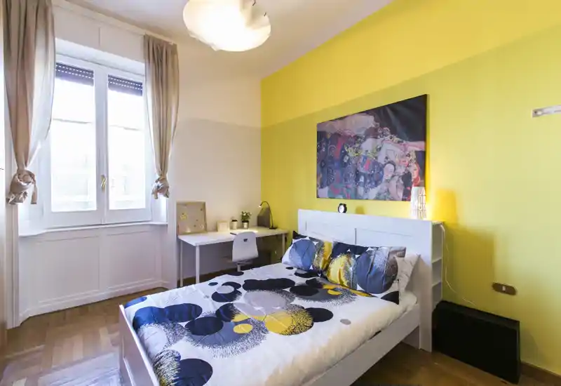 Camera da letto luminosa e curata, con letto matrimoniale, cuscini decorativi e ampia finestra che garantisce luce naturale; scrivania accanto alla finestra adatta per lavoro o studio.