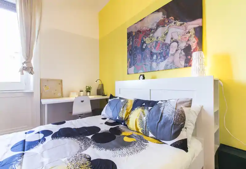 Camera da letto luminosa e curata con letto matrimoniale, testata bianca con scaffalatura integrata, scrivania vicino alla finestra e quadro colorato che dona un’atmosfera moderna e accogliente.