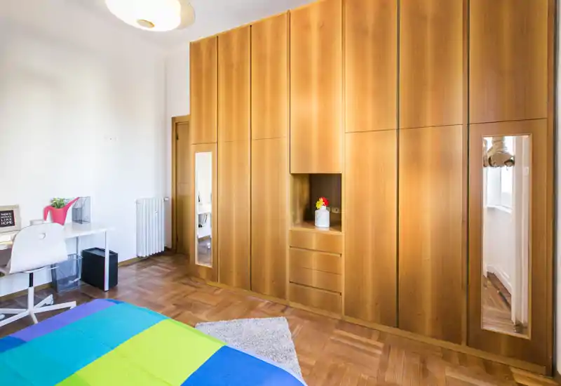 Camera da letto luminosa e ordinata con grande armadio in legno, pavimento in parquet e una coperta colorata. La stanza include una scrivania, specchi sulle ante e luce naturale gradevole.