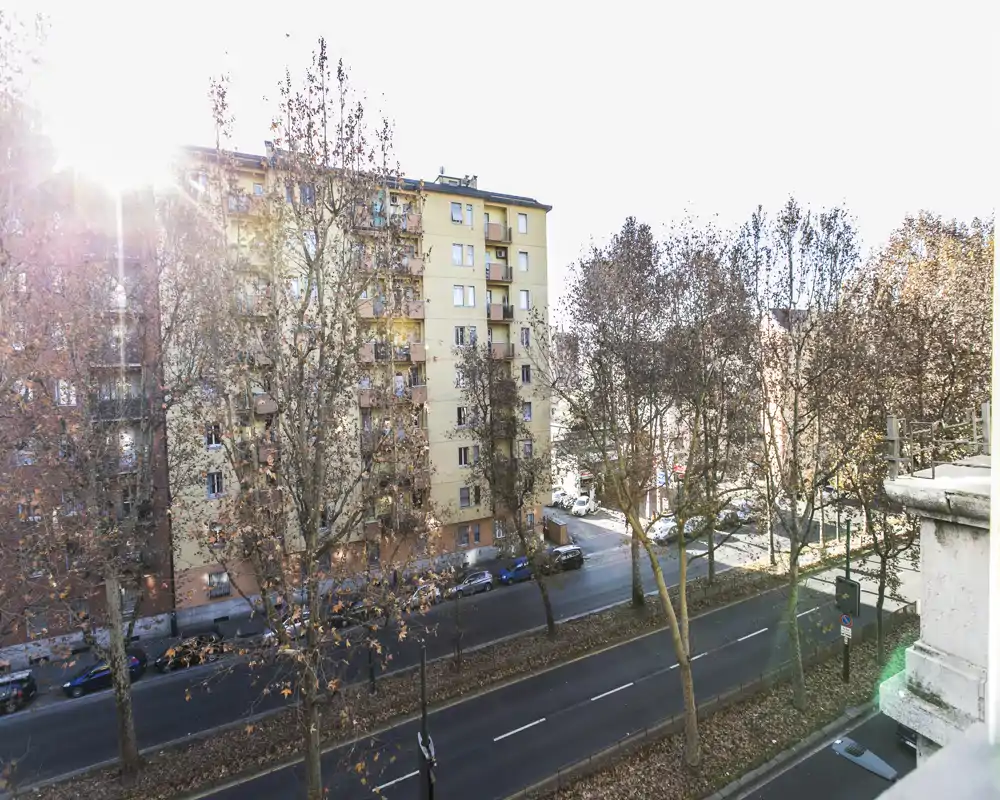 Vista su strada e palazzi da un balcone o finestra con alberi e luce diurna; utile per mostrare il quartiere e gli edifici vicini.
