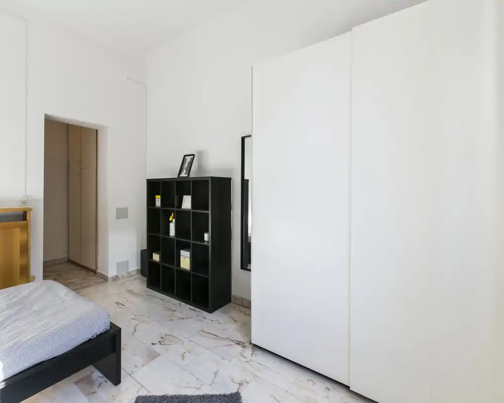 Camera da letto luminosa e minimale con letto visibile, grande armadio bianco e libreria nera; pavimento effetto marmo e arredo moderno essenziale.