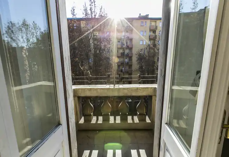 Piccolo balcone che si affaccia su un cortile cittadino soleggiato, con ringhiera in pietra decorativa e palazzina sullo sfondo.