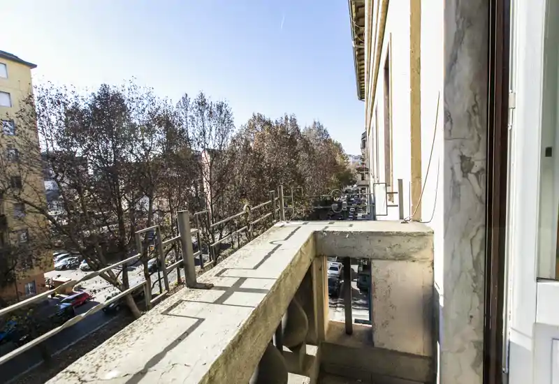 Balcone soleggiato e stretto con ringhiera in metallo e balaustra in pietra che si affaccia su una via alberata — perfetto per un caffè mattutino.