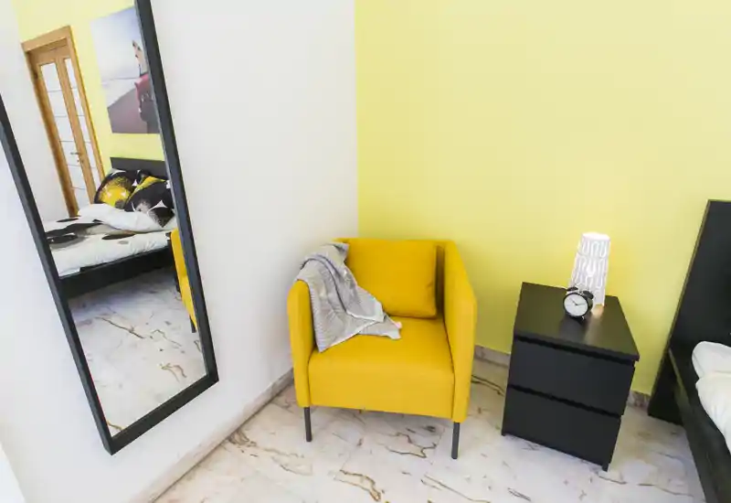 Angolo della camera luminoso e moderno con una poltrona gialla, comodino con lampada e sveglia, e parte del letto visibile sul bordo dell’immagine e nello specchio.