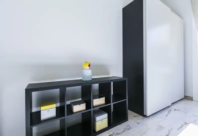 Angolo di soggiorno minimal con scaffalatura modulare e un armadio alto; luce naturale intensa e scatole decorative con un vaso donano un senso pulito e moderno.