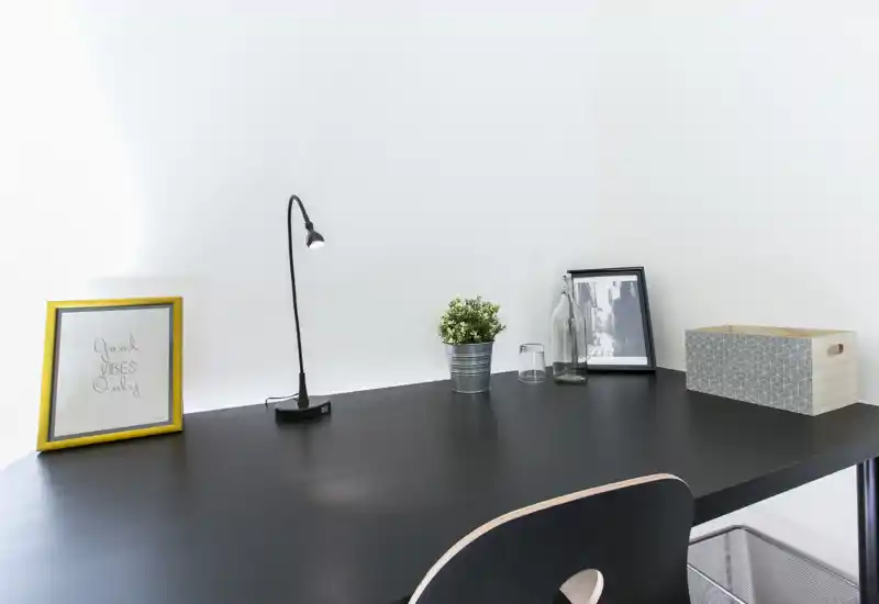 Area di lavoro luminosa e minimale con piano nero, lampada da tavolo, piccoli oggetti decorativi e cornici: uno spazio ordinato e ben illuminato per studiare o lavorare.