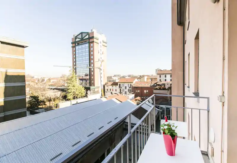 Balcone privato soleggiato con un piccolo tavolo e una pianta, con vista sui tetti della città e su un edificio moderno — perfetto per il caffè mattutino.