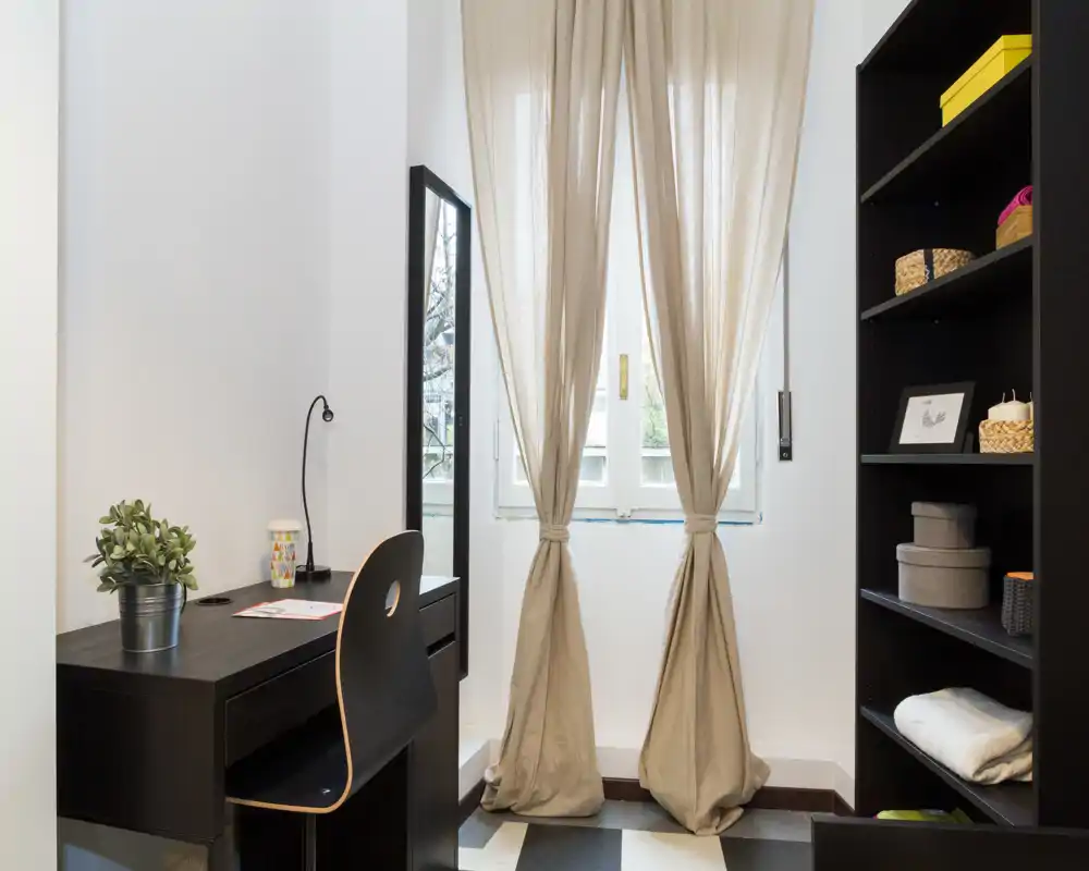 Piccola stanza luminosa con scrivania e scaffale vicino alla finestra con tende leggere; ideale come angolo studio o workspace in appartamento.