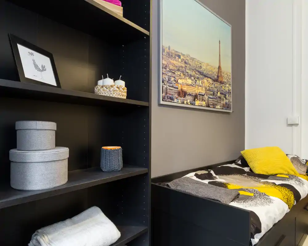 Camera da letto compatta e curata con un letto singolo rivestito da biancheria a fantasia e un cuscino giallo; scaffalatura con scatole decorative che aggiunge spazio e carattere.