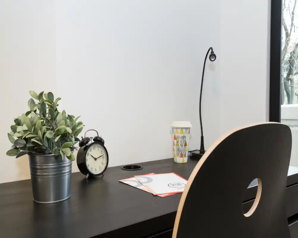 Angolo scrivania ordinato con piano nero moderno, lampada, sveglia e pianta in vaso — un compatto angolo studio con luce naturale.