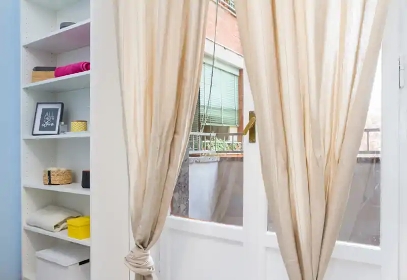 Pequeño balcón visible a través de puertas de cristal con cortinas beige claro; estantería ordenada y luz natural suave que crean una transición tranquila hacia el exterior.