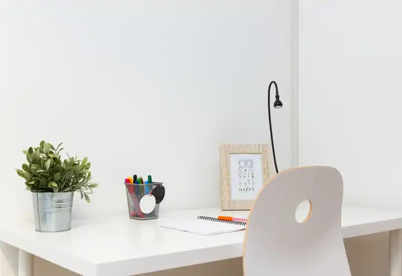 Rincón de oficina en casa minimalista con escritorio blanco, silla, lámpara, marco, portalápices y planta en maceta; espacio de trabajo luminoso y ordenado.