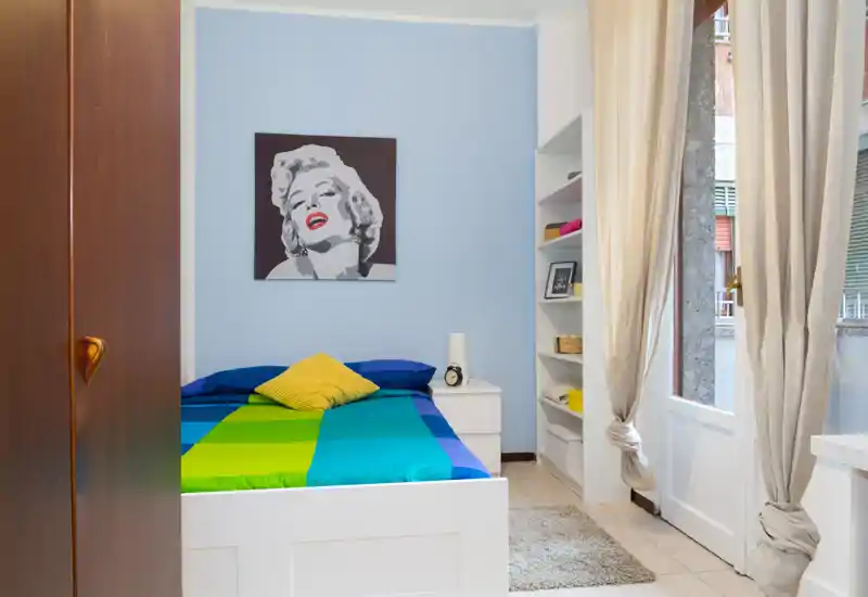 Dormitorio luminoso y bien presentado con colcha colorida, cojines decorativos y un cuadro pop-art; luz natural entra por la puerta con cortinas.