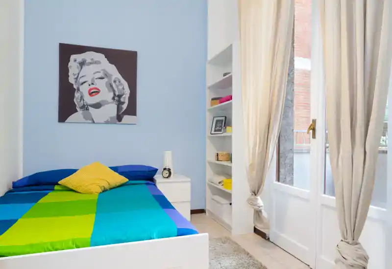 Dormitorio moderno y luminoso con una cama doble colorida, mesita de noche y estanterías junto a una gran ventana/puerta que aporta luz natural.