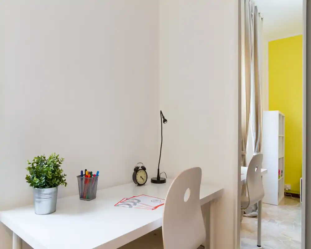 Zona de trabajo luminosa y minimalista con escritorio blanco, silla, lámpara de mesa, planta pequeña y material de escritura — perfecto para estudiar o trabajar desde casa.