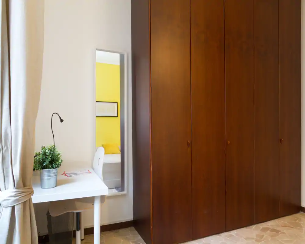 Rincón de dormitorio con un gran armario de madera y un pequeño escritorio; la cama solo se ve reflejada en el espejo alto. La luz natural y la decoración neutra mantienen el espacio luminoso.
