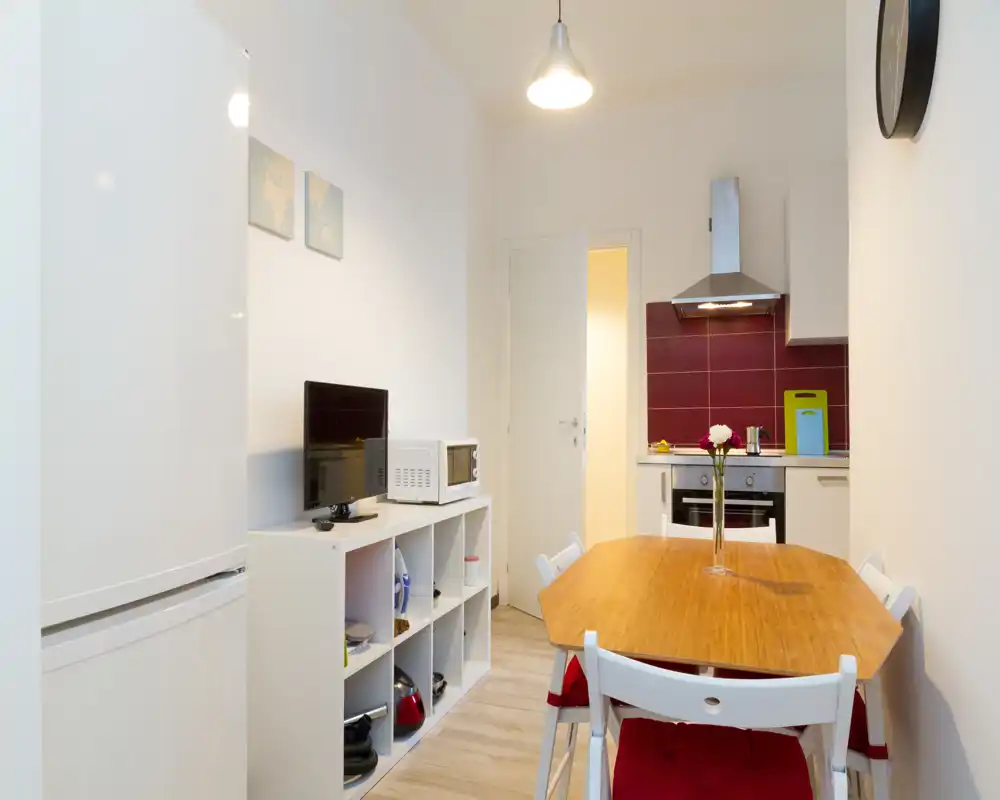 Cocina compacta abierta con mesa de comedor de madera para cuatro. Distribución ordenada con electrodomésticos modernos, centro de mesa decorativo y paredes claras que realzan la luz.