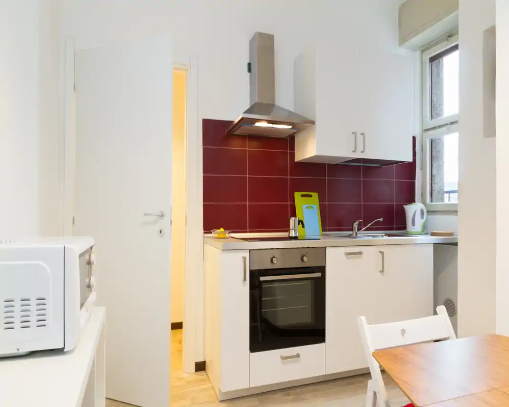 Cocina compacta y moderna con horno, placa y fregadero, salpicadero de azulejos rojos y pequeña mesa — luminosa y ordenada, ideal para un estudio.