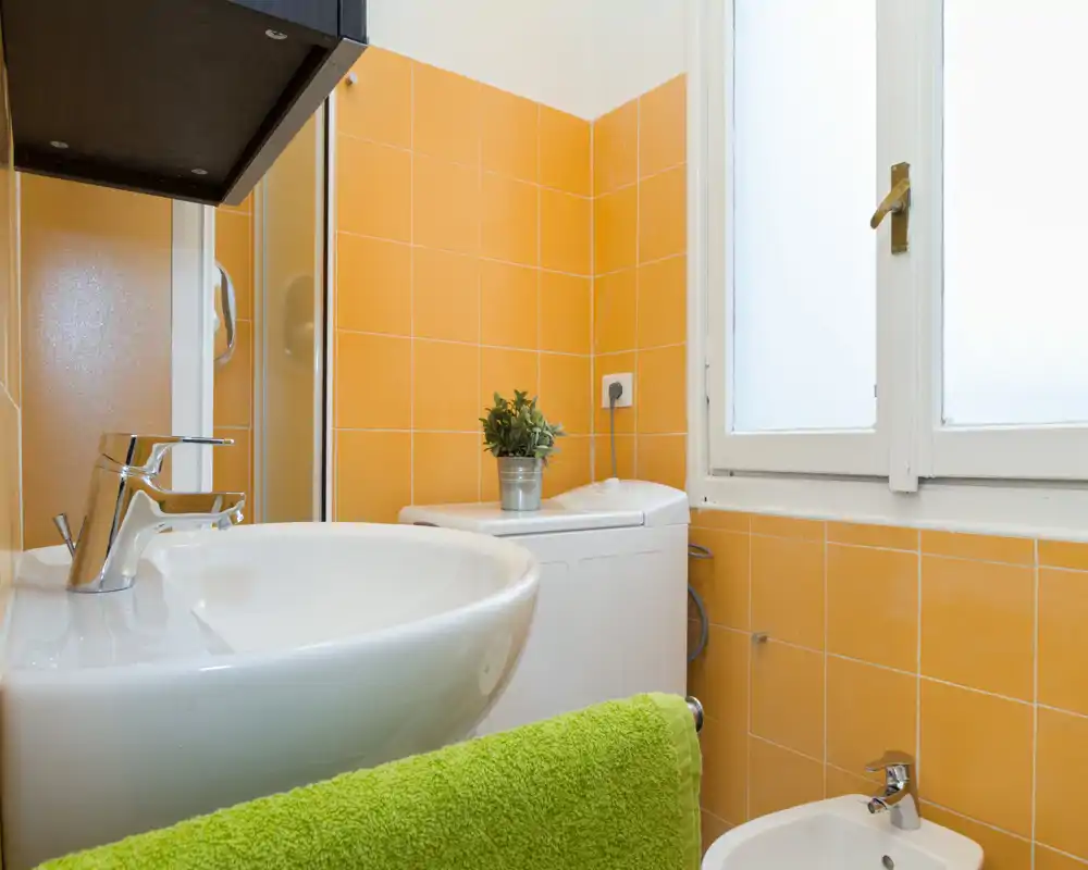 Baño compacto con azulejos naranjas, lavabo y bidé blancos, luz natural por una ventana translúcida y una pequeña lavadora; una toalla verde aporta color.