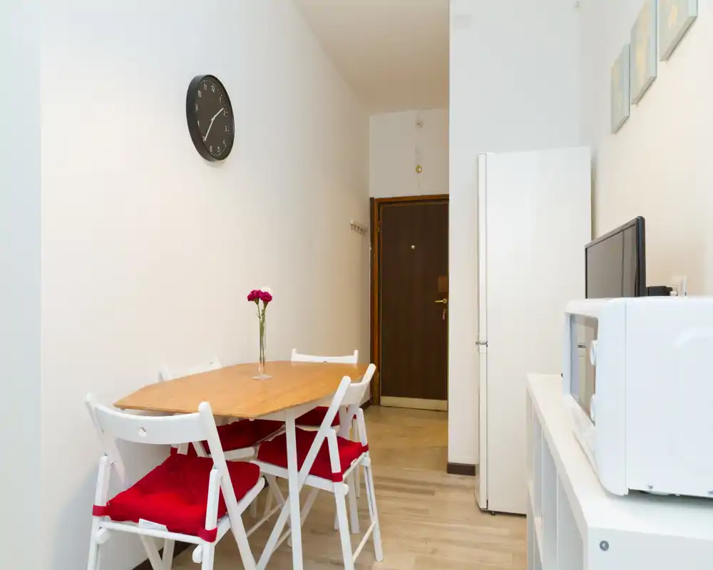 Zona de comedor luminosa y ordenada con mesa de madera, cuatro sillas plegables blancas con cojines rojos y un jarrón como centro de mesa — ideal para apartamentos compactos.