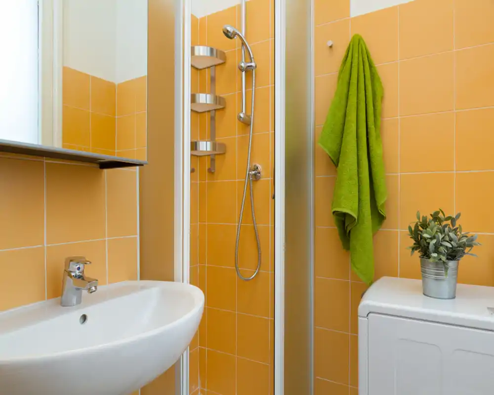 Baño compacto y luminoso con azulejos naranjas, ducha en esquina con puertas de cristal, lavabo blanco y una toalla colgada en la pared.