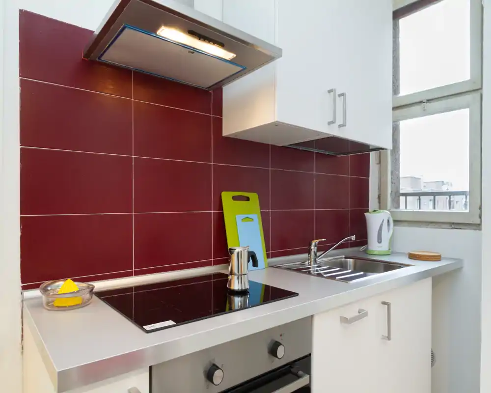 Cocina moderna y compacta con placa de inducción brillante, fregadero de acero, campana extractora y salpicadero de azulejos rojo; encimera ordenada con hervidor y tabla de cortar junto a la ventana.